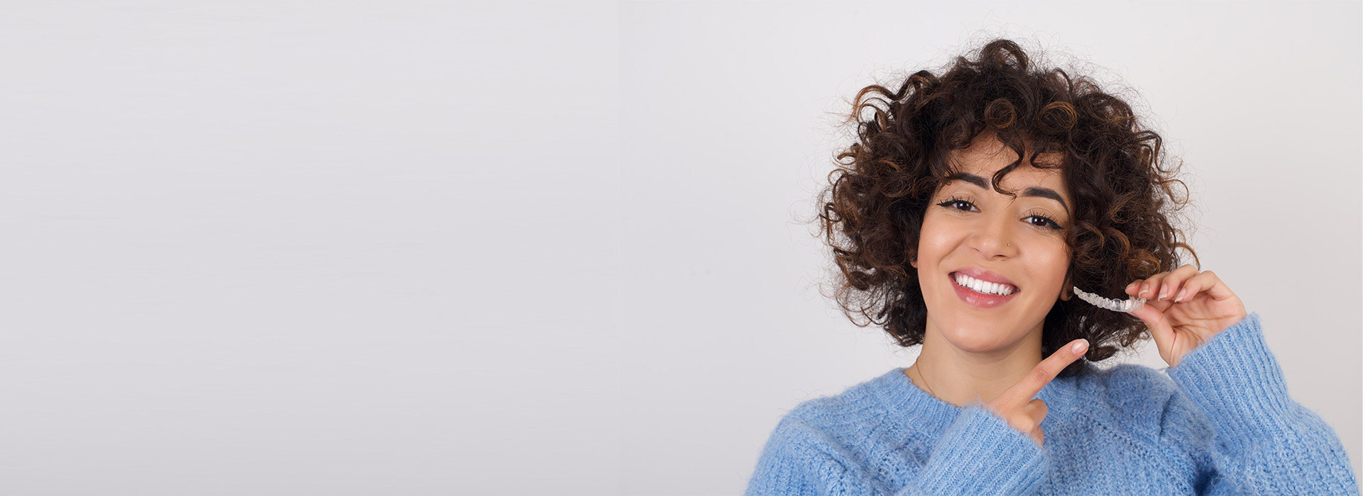 This is a color photograph featuring a person smiling at the camera with their hand near their face. They have curly hair, are wearing a blue top, and appear to be indoors against a plain background.