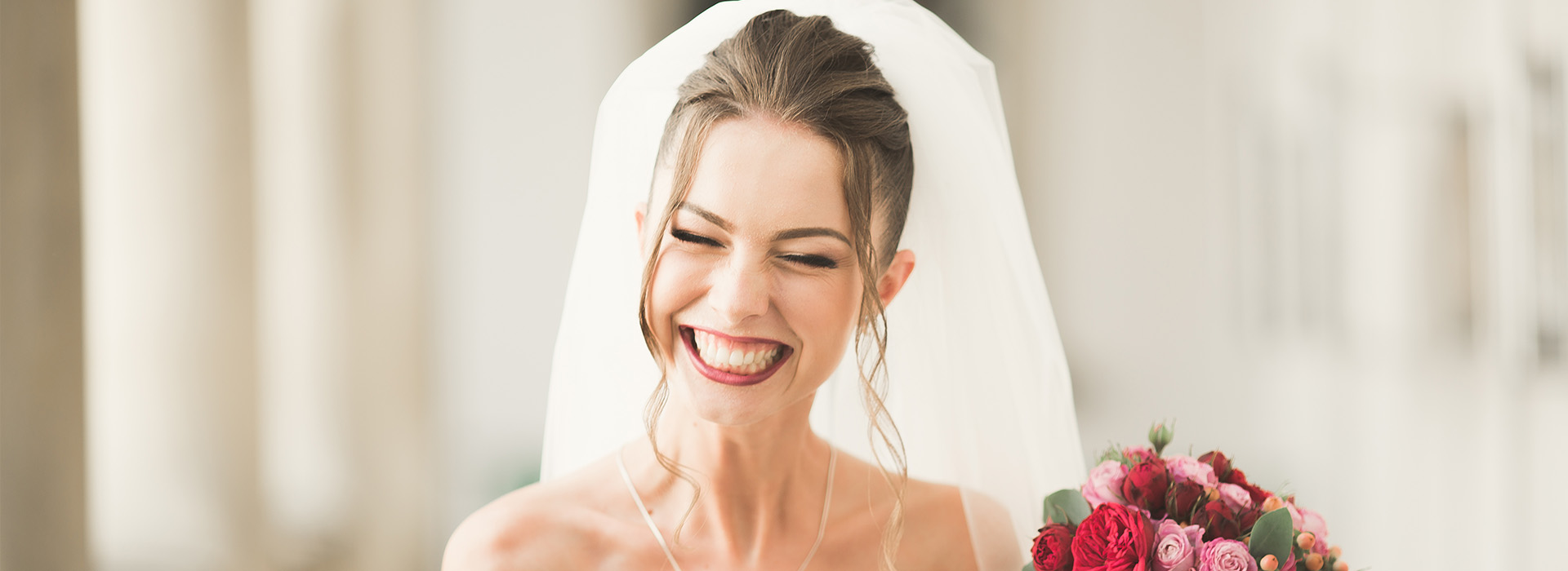 A bride with a radiant smile wearing a white wedding dress and veil, with her face visible in the foreground, against a blurred background of an indoor setting.