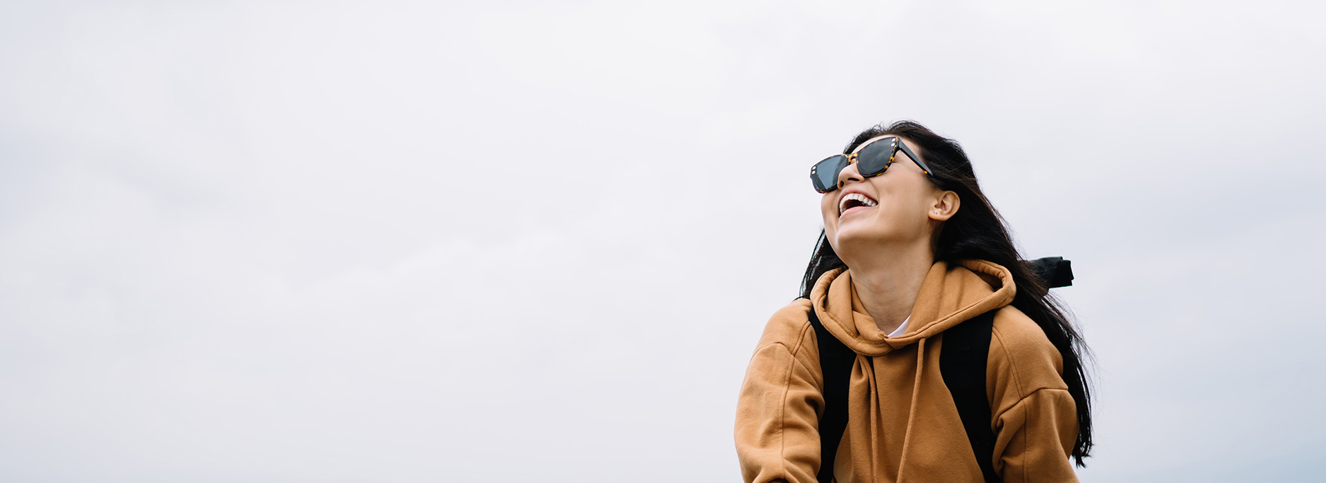 A person with sunglasses on their head is smiling at the camera while standing outdoors against a cloudy sky, wearing a backpack and a dark-colored jacket.