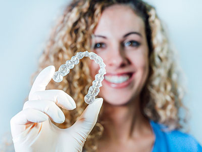 A woman holding up a clear plastic tray containing a single white pearl-shaped object, likely a piece of jewelry or an item for sale, against a blurred background that suggests a store setting.