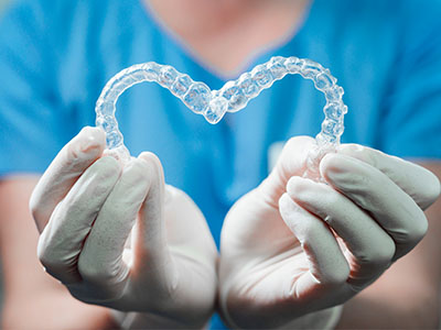The image shows a person s hands holding a heart-shaped object made of clear plastic material, which appears to be a dental appliance, against a blue background that suggests a medical setting.