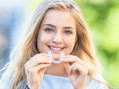 The image shows a young woman with light hair smiling at the camera while holding up a clear plastic dental retainer with her teeth.