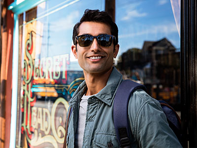 A man wearing sunglasses and a backpack stands outside a storefront, smiling at the camera.