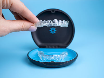 The image shows a person s hand holding an open case containing a set of clear dental aligners with the brand name  Invisalign  visible on the packaging, placed against a blue background.
