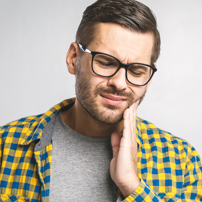 A man with glasses, beard, and mustache, wearing a yellow plaid shirt, holding his hand on his chin, with a look of concern or worry on his face.